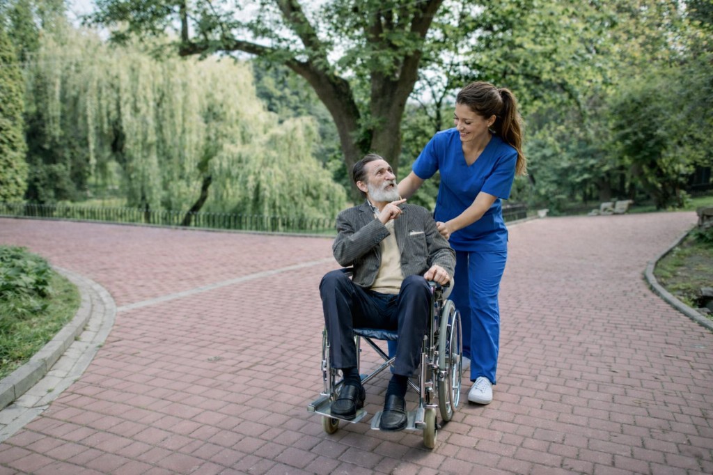 A caregiver in blue scrubs pushing an older man in a wheelchair along a sunny park path, both engaged in a friendly conversation.