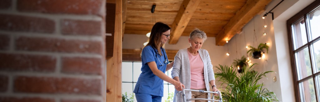 A caregiver in blue scrubs supporting an older woman as she walks with a walker in a warm, well-lit indoor setting.